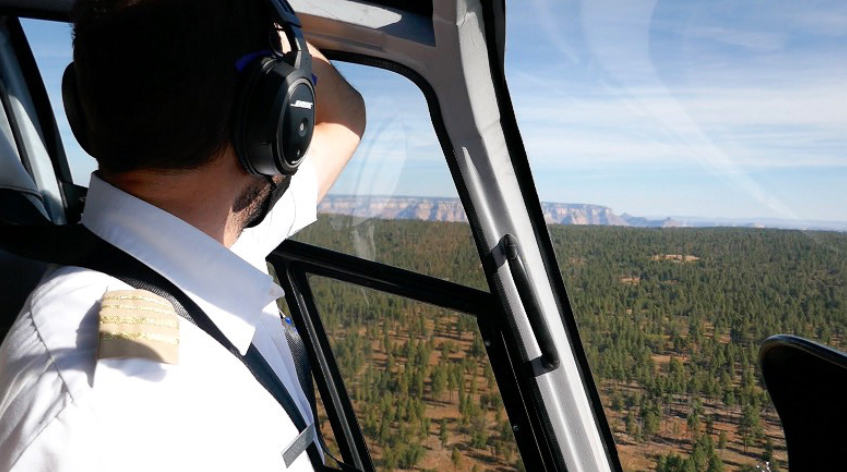 back profile of a pilot looking out the helicopter window towards the grand canyon on a clear day with forest below