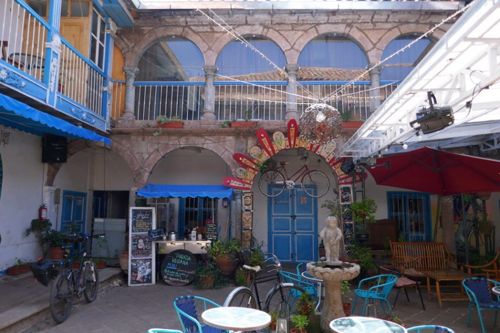 colorful courtyard outside of cusco culinary with open air balconies, bicycles, a cafe bar, a fountain with a small statue, stone arches and blue accent paint