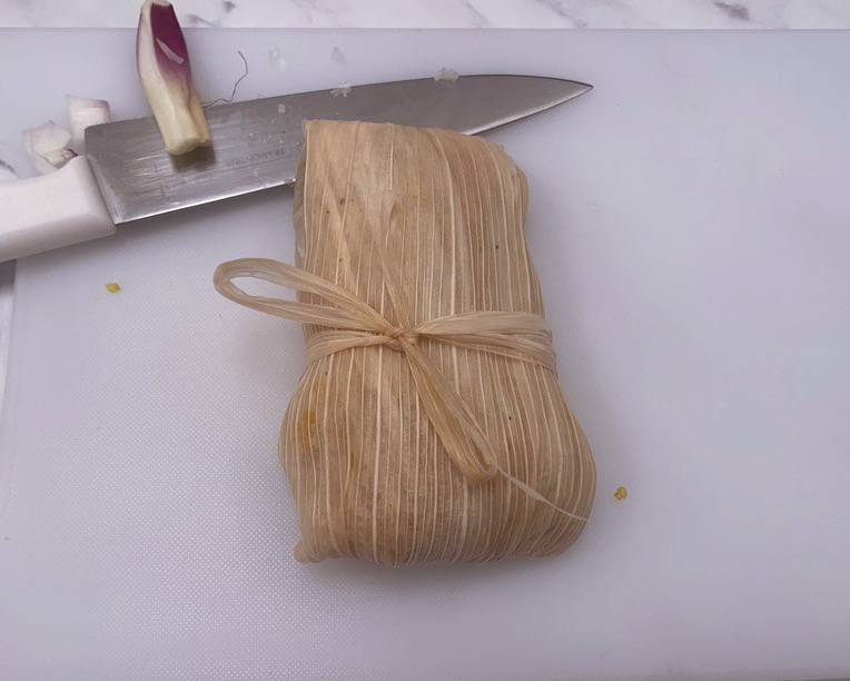 a tomale tied into a bow before being cooked, sitting on a white table top with a knife behind it