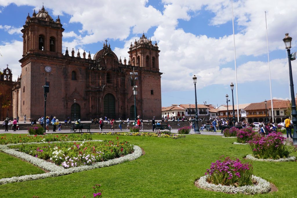 the cusco cathedral on a sunny day with lush green grass and pink flowers in the garden square in front of it in Cusco's town center