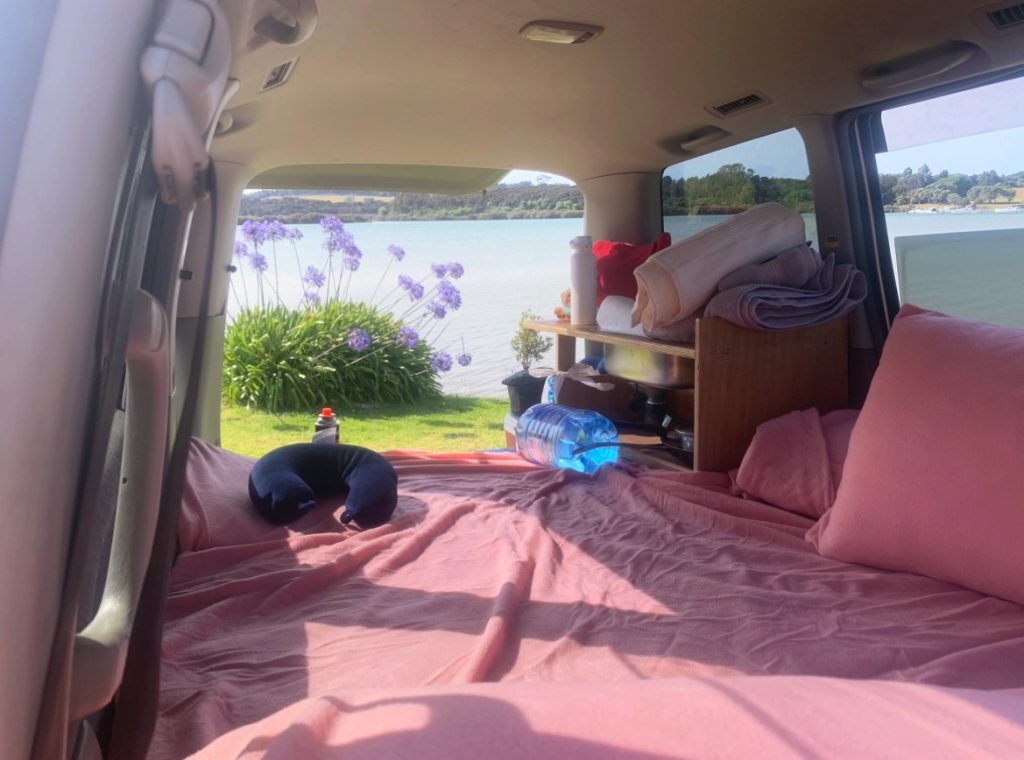 inside of the sleeping space in a converted mini van with pink fitted sheets in front of a lake in new zealand